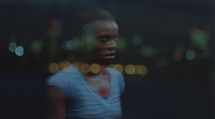 A woman watches from the night, lights reflected unfocused from a window