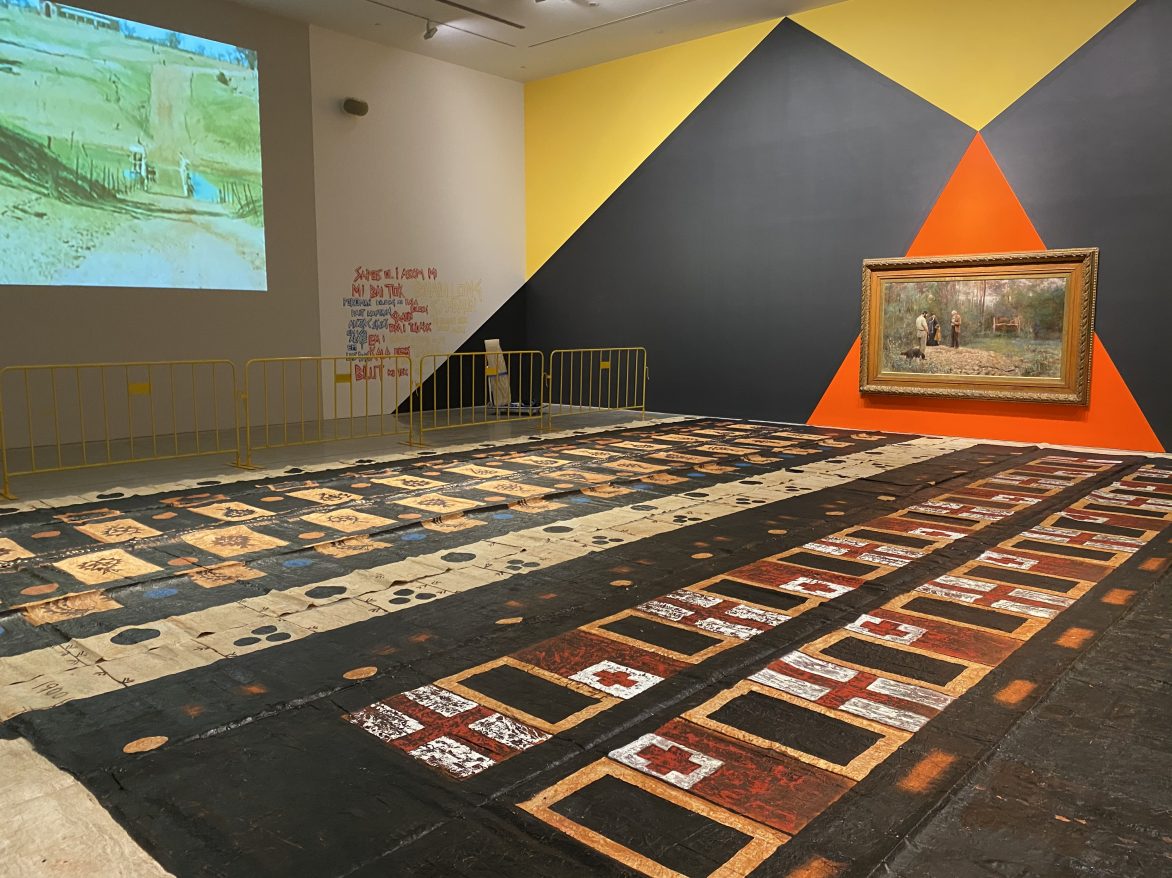 Exhibition view, NIRIN – the 22nd Biennale of Sydney, 2020, Museum of Contemporary Art, Sydney, Australia.
Artworks in view:
Eric Bridgeman, Rot Bung (Junction), 2019-20. Acrylic on wall, dimensions variable. Courtesy of the artist.
Frederick McCubbin, A bush burial, 1890. Oil on canvas, 122.5 x 224.5cm. Courtesy of Geelong Gallery.
Kulimoe’anga Stone Maka, Kuini Haati 2 (Two Queen Heart), 2008–10, and Togo mo Bolataane (Tonga and Britain), 2008–10. Oil, clay, daye on tapa cloth. Both 427 x 1115 cm. Courtesy of the artist.
H.R. Balfourm Dendroglyphs of the Kalimangle Bora Ground, 1949. Single channel digital video from 16mm, colour, silent, 53 mins. Courtesy the Australian Institute of Aboriginal and Torres Strait Islander Studies.
Brook Andrew, Powerful Object: Dendroglyph, 2020.3D model printed in clear filament, 109.5 x 49 x 23.5cm. Model made from a dendroglyph tree section, site marker, 19th Century. Collection of the Pitt Rivers Museum, University of Oxford [1948.12.77].